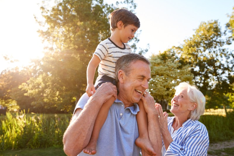 Grandparents Giving Grandson Ride On Shoulders In Summer Park Against Flaring Sun