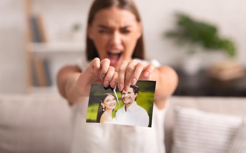 Breakup. Furious Woman Ripping Wedding Photo With Ex-Husband Sitting On Sofa Indoor After Divorce. Selective Focus