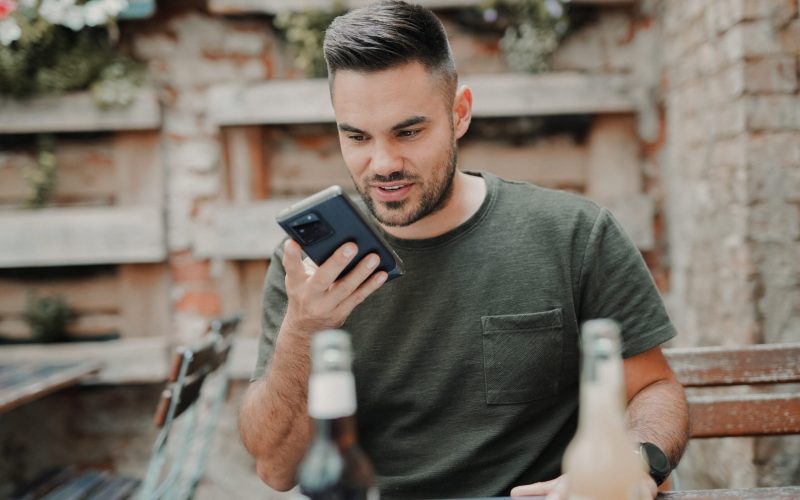 Man talking into the phone sending a voice message, sitting in a caffee work and travelling with a smartwatch, two drinks and leisure cloth. He is communicating via his smartphone.