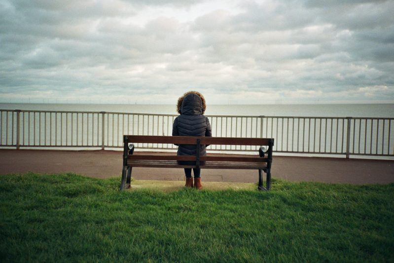 A figure with a Winter coat on sitting on a park bench looking out to sea on a grey overcast Winter day. Shot on film.
