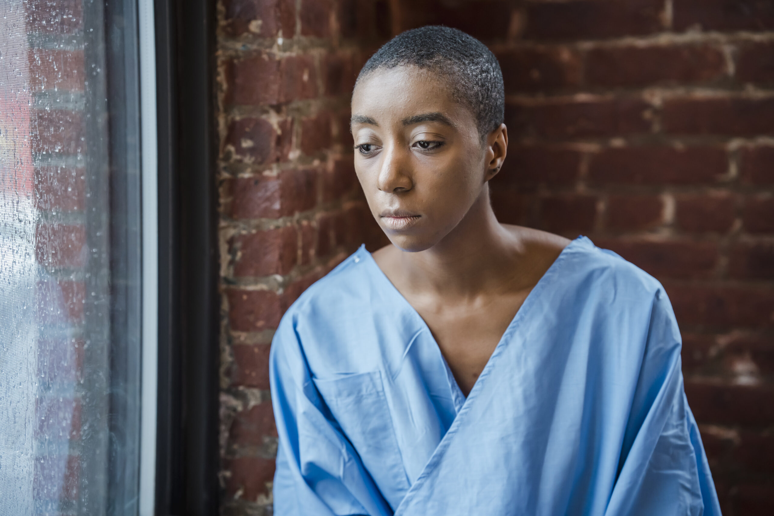 Melancholy black woman in medical clothes sitting near window against brick wall