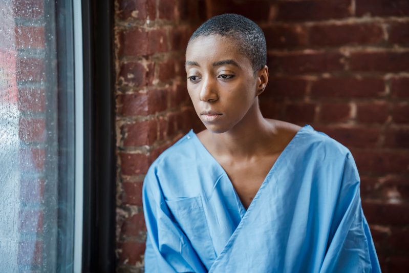 Melancholy black woman in medical clothes sitting near window against brick wall