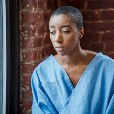 Melancholy black woman in medical clothes sitting near window against brick wall